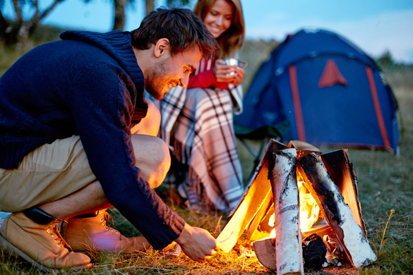 Young-man-kindling-firewood-in-the-countryside-with-girl-and-tent