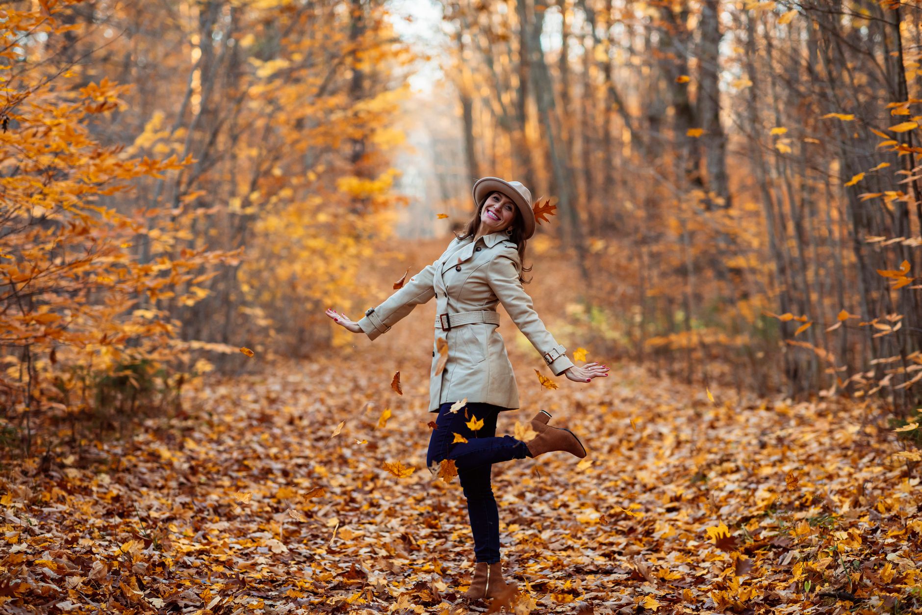 A woman joyfully poses among falling autumn leaves in a forest, embracing the vibrant fall season.