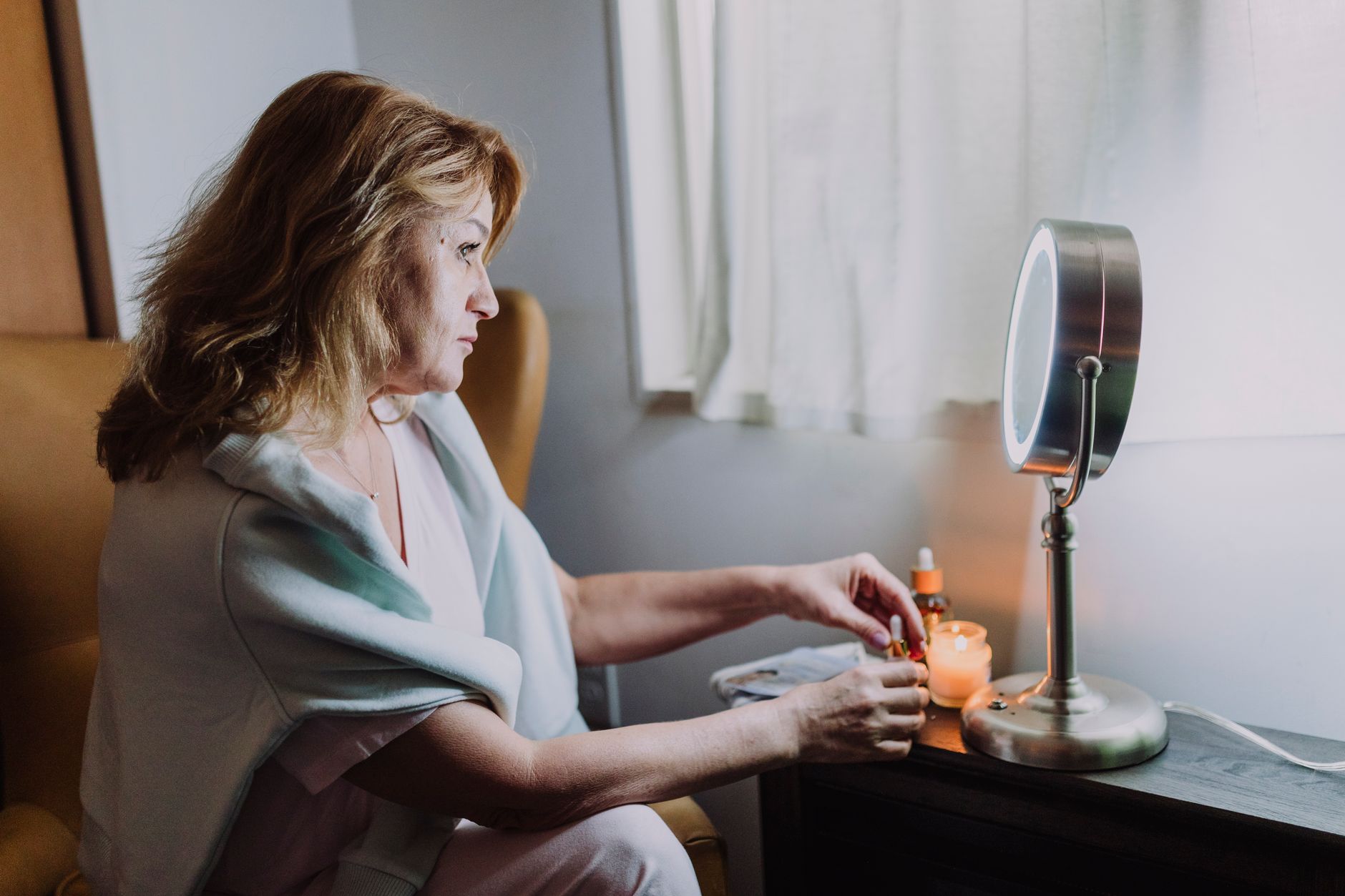 A woman enjoys a peaceful skincare routine with candles by a window.