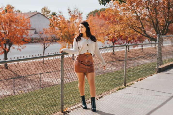 Fashionable woman poses by a fence amidst colorful fall foliage during the day.