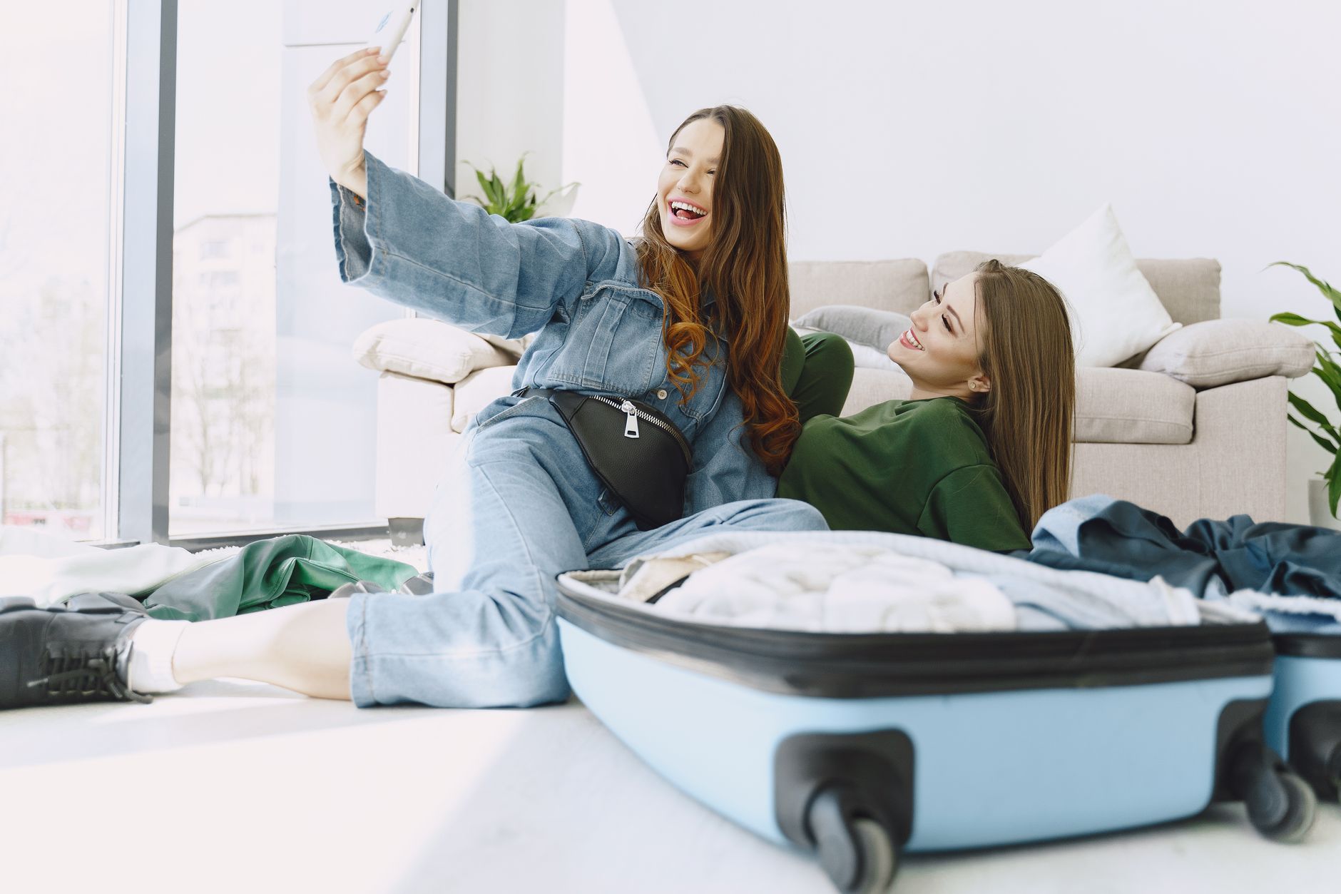 Happy young female friends in trendy outfits having fun and taking selfie while packing suitcase in cozy room