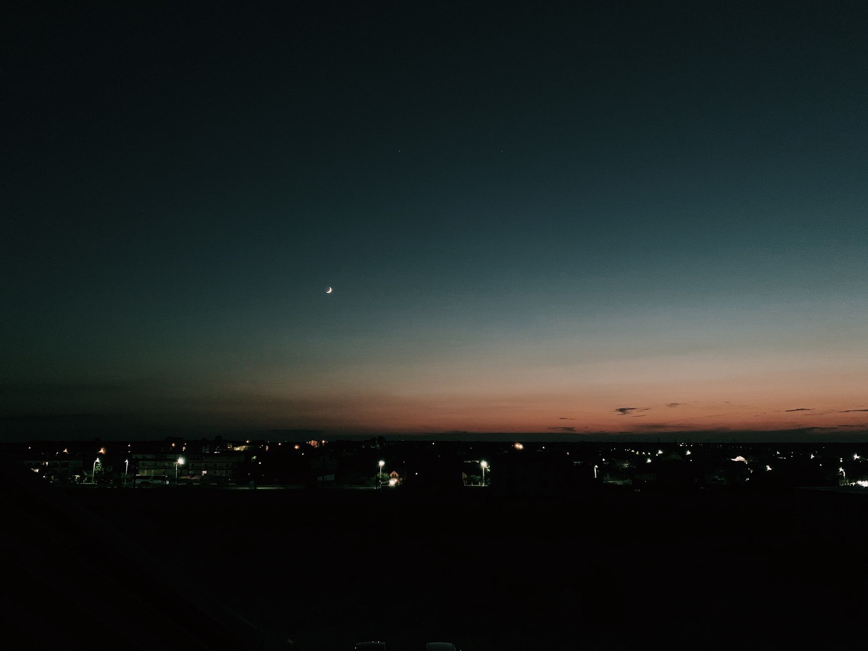 Starless dark sky with new moon glowing over small settlement houses illuminated by electric lights