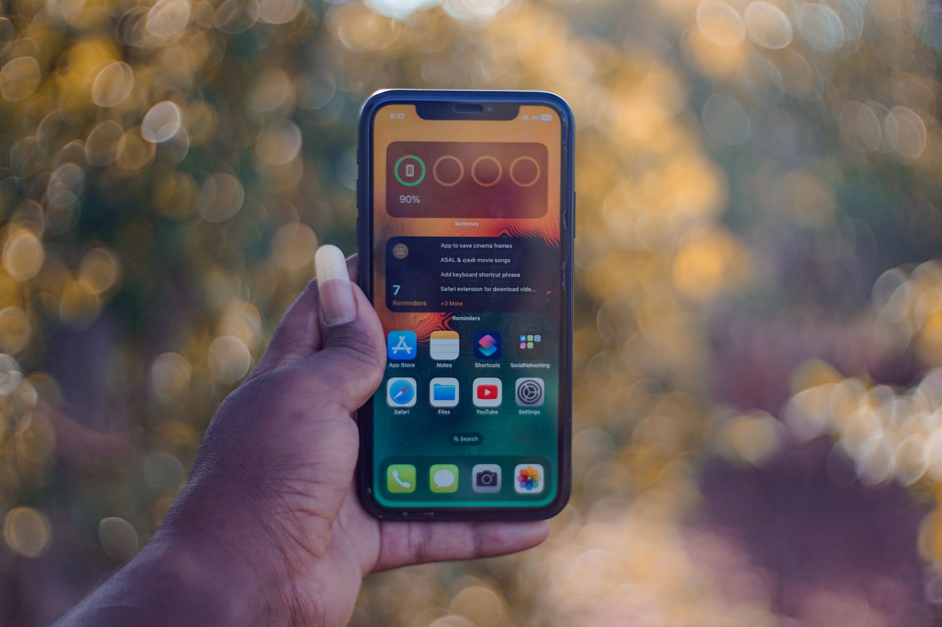 A hand holding a smartphone showing home screen apps, outdoors with bokeh background.