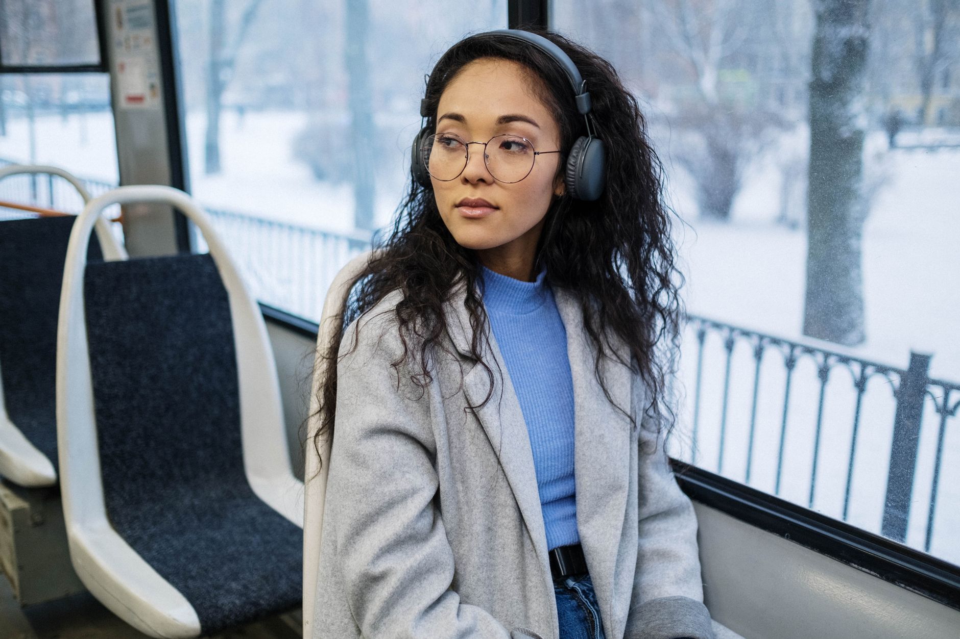 Stylish woman in gray coat enjoying music through wireless headphones on a snowy day.