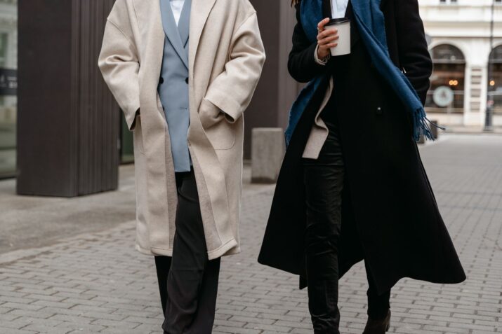 Two adults in stylish coats walk on a city street, enjoying a coffee break.