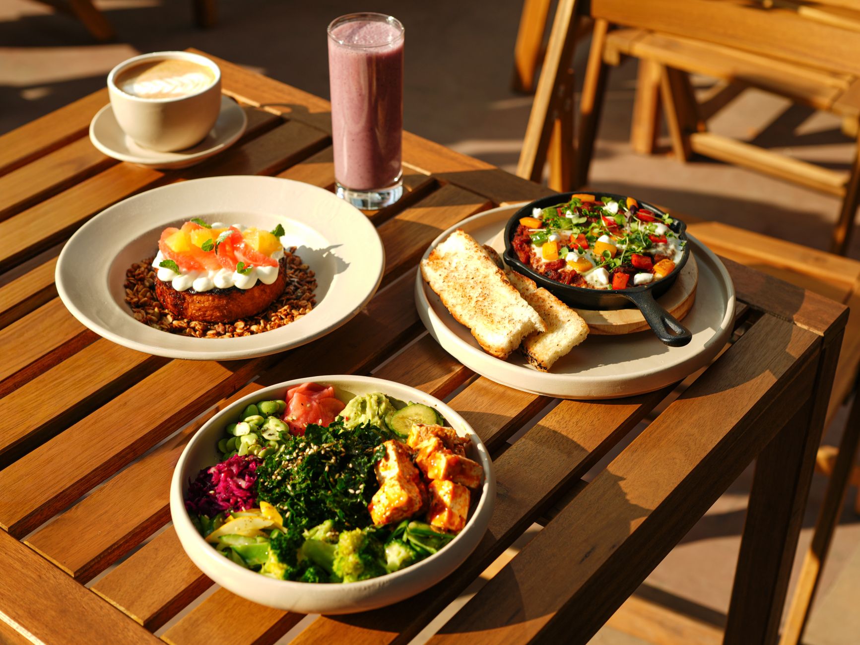 A vibrant set of healthy dishes including a smoothie and coffee on an outdoor wooden table.