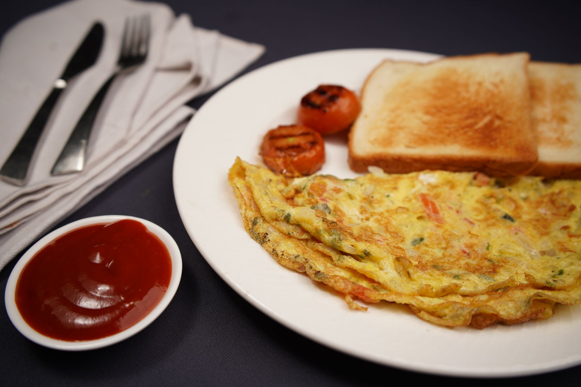 A hearty breakfast featuring omelette, toast, grilled tomatoes, and ketchup on a black background.