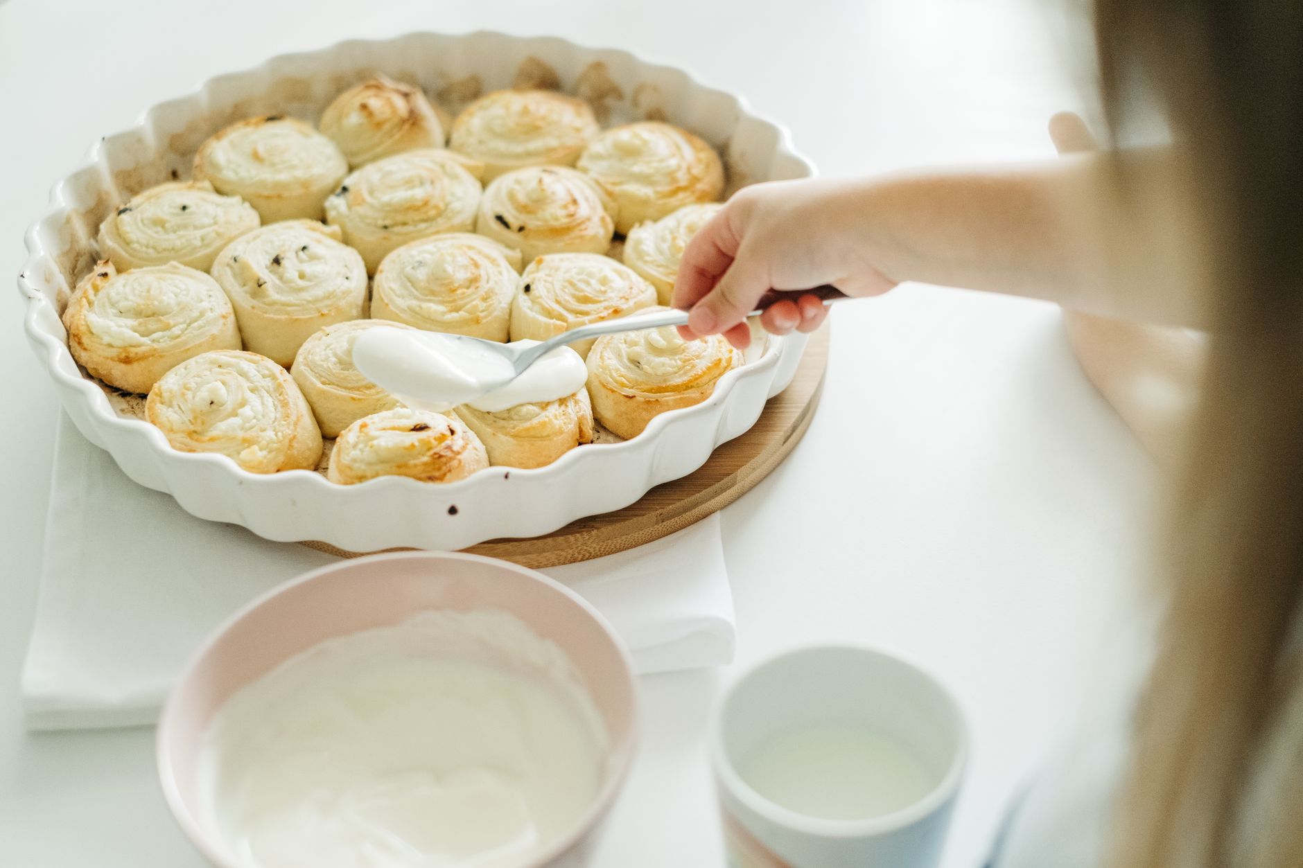 A child spreads creamy icing over freshly baked cinnamon rolls, highlighting a cozy food preparation scene.