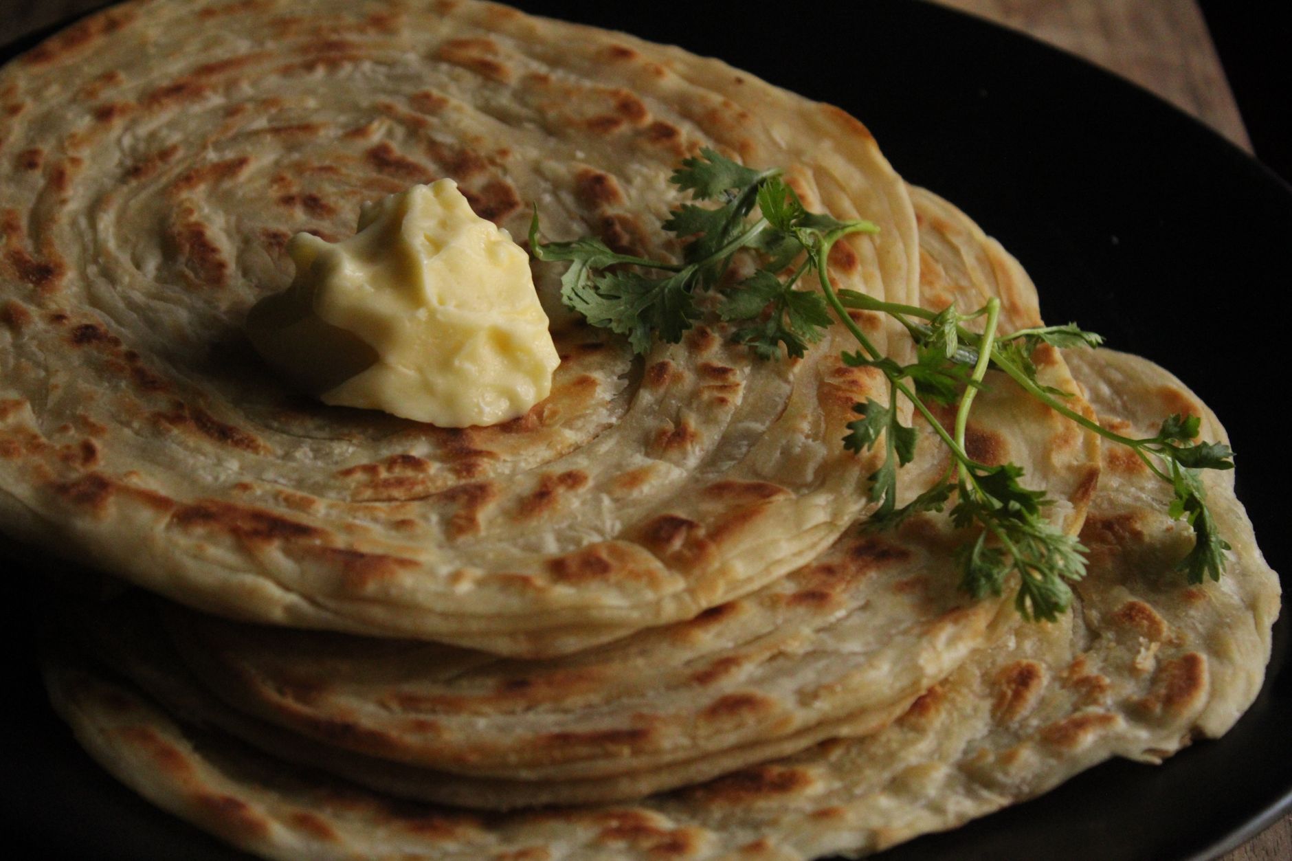 Delicious flaky parathas with butter and cilantro garnish on a black plate.