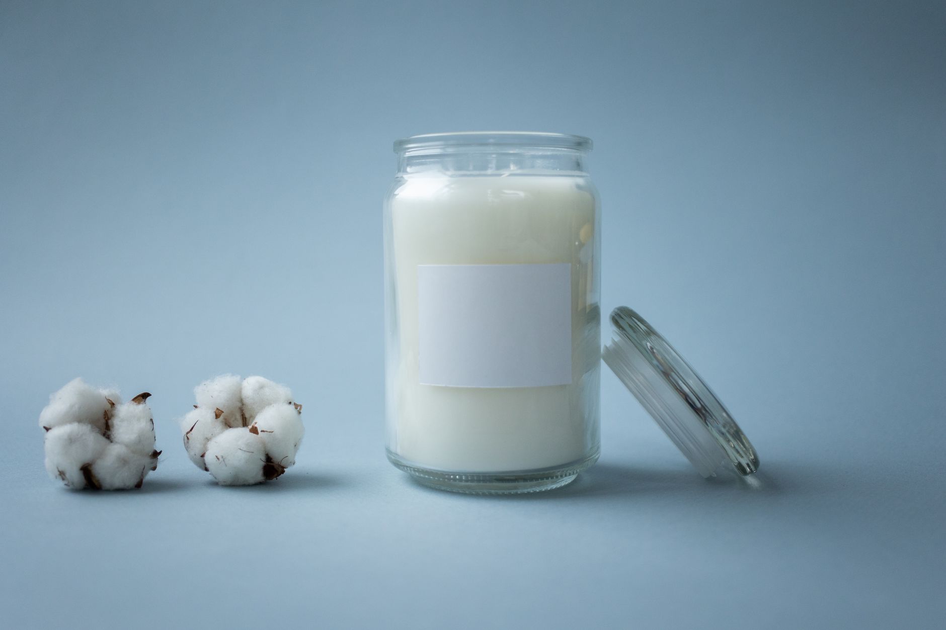 A minimalist shot of a glass jar candle beside cotton balls on a soft blue backdrop.