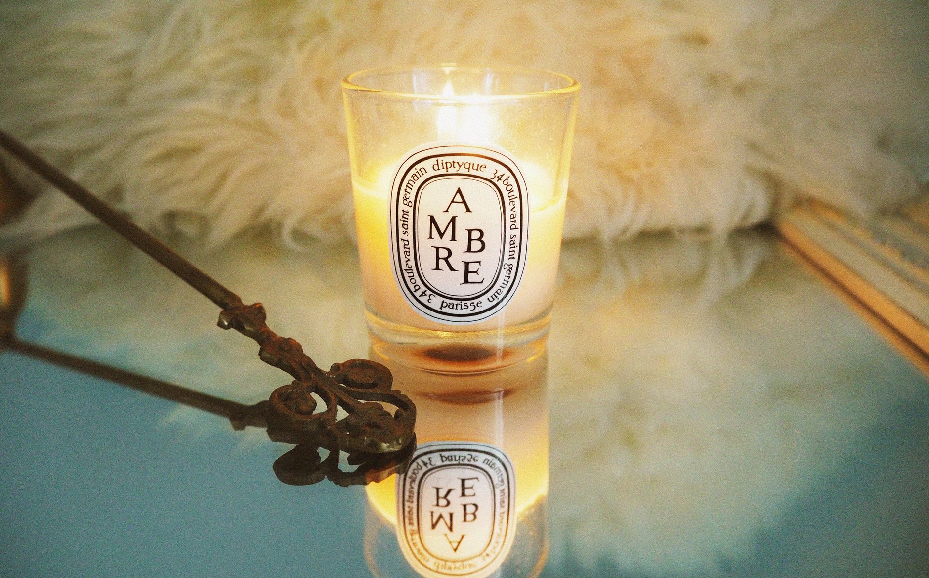 A cozy scene featuring a Diptyque candle reflecting warmly against a mirrored surface with soft lighting.