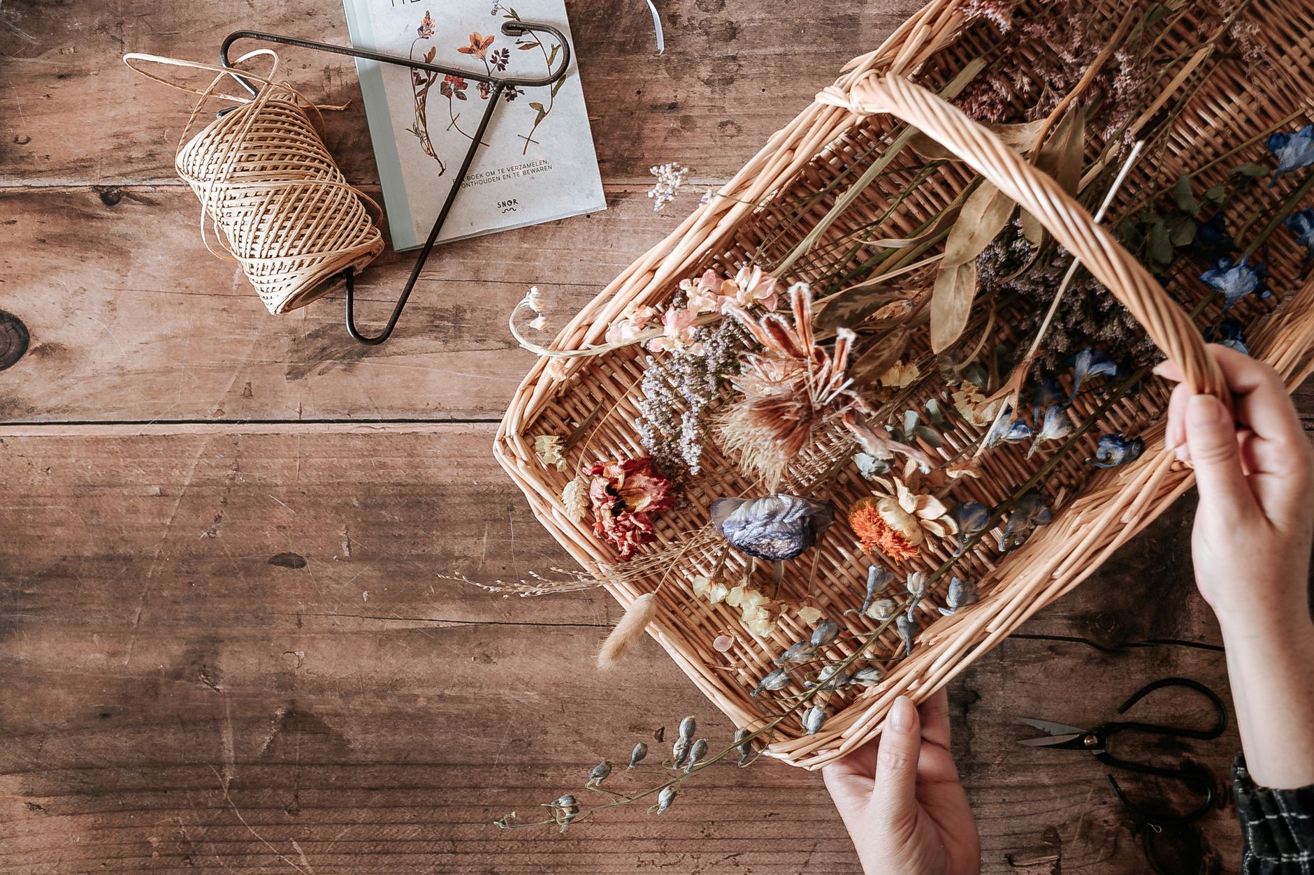 Hands arranging a variety of dried flowers in a wicker basket on a rustic wooden surface.