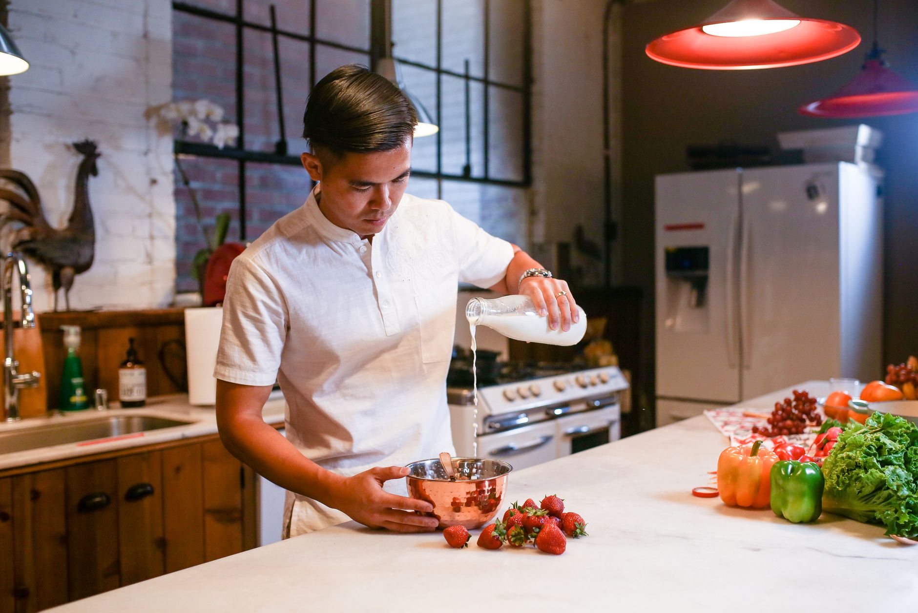 A man pouring milk into a copper bowl on a kitchen counter filled with fresh produce.