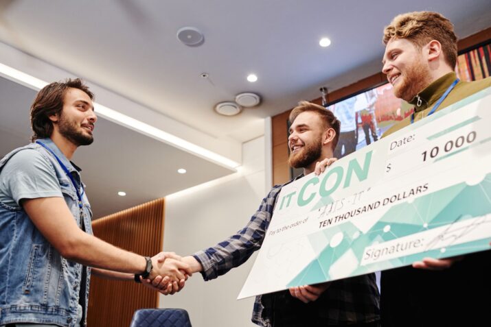 Two men celebrating with a $10,000 check at an indoor event, shaking hands and smiling.