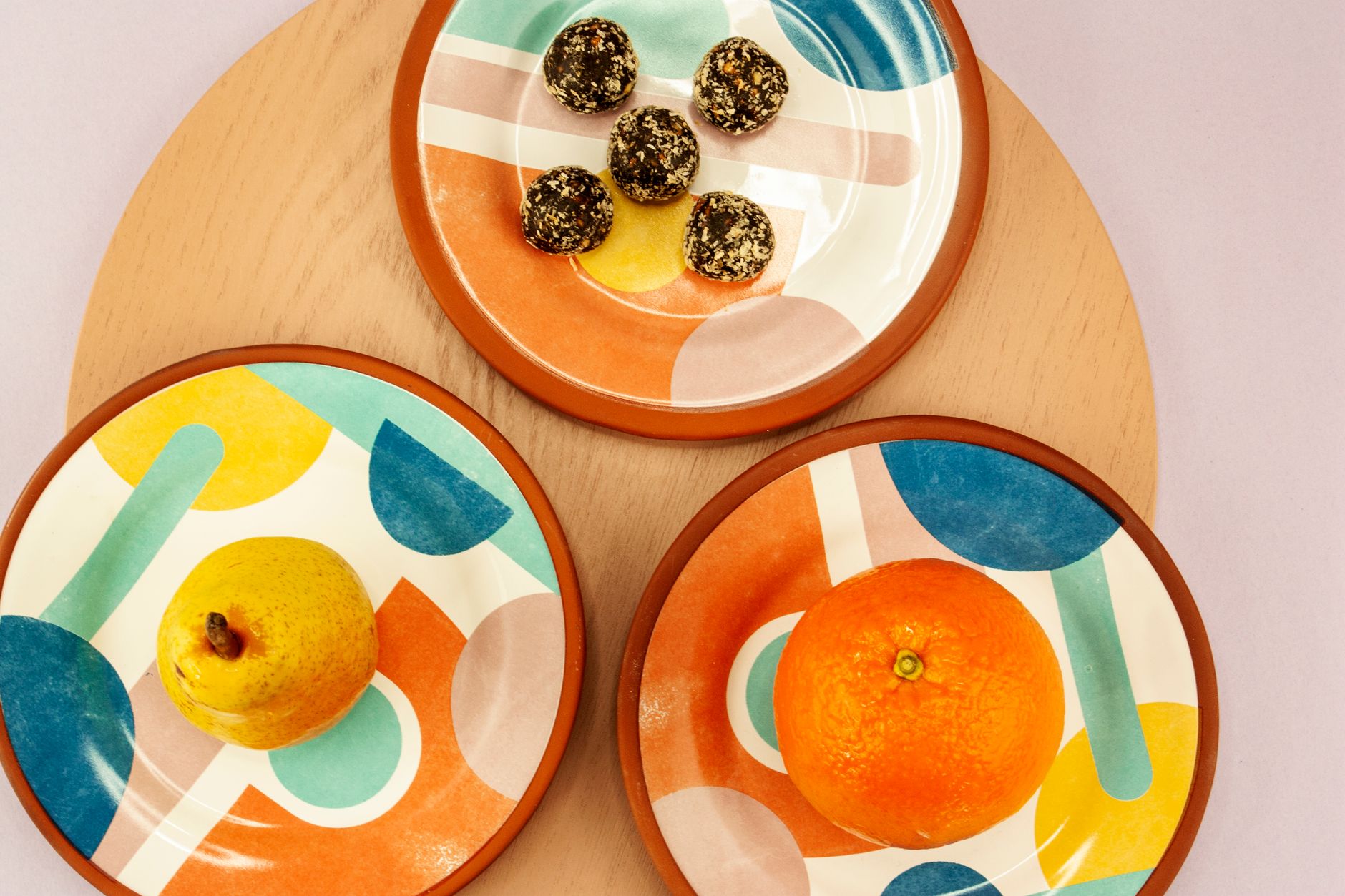 Vibrant geometric plates showcasing a pear, an orange, and snacks on a wooden tray.