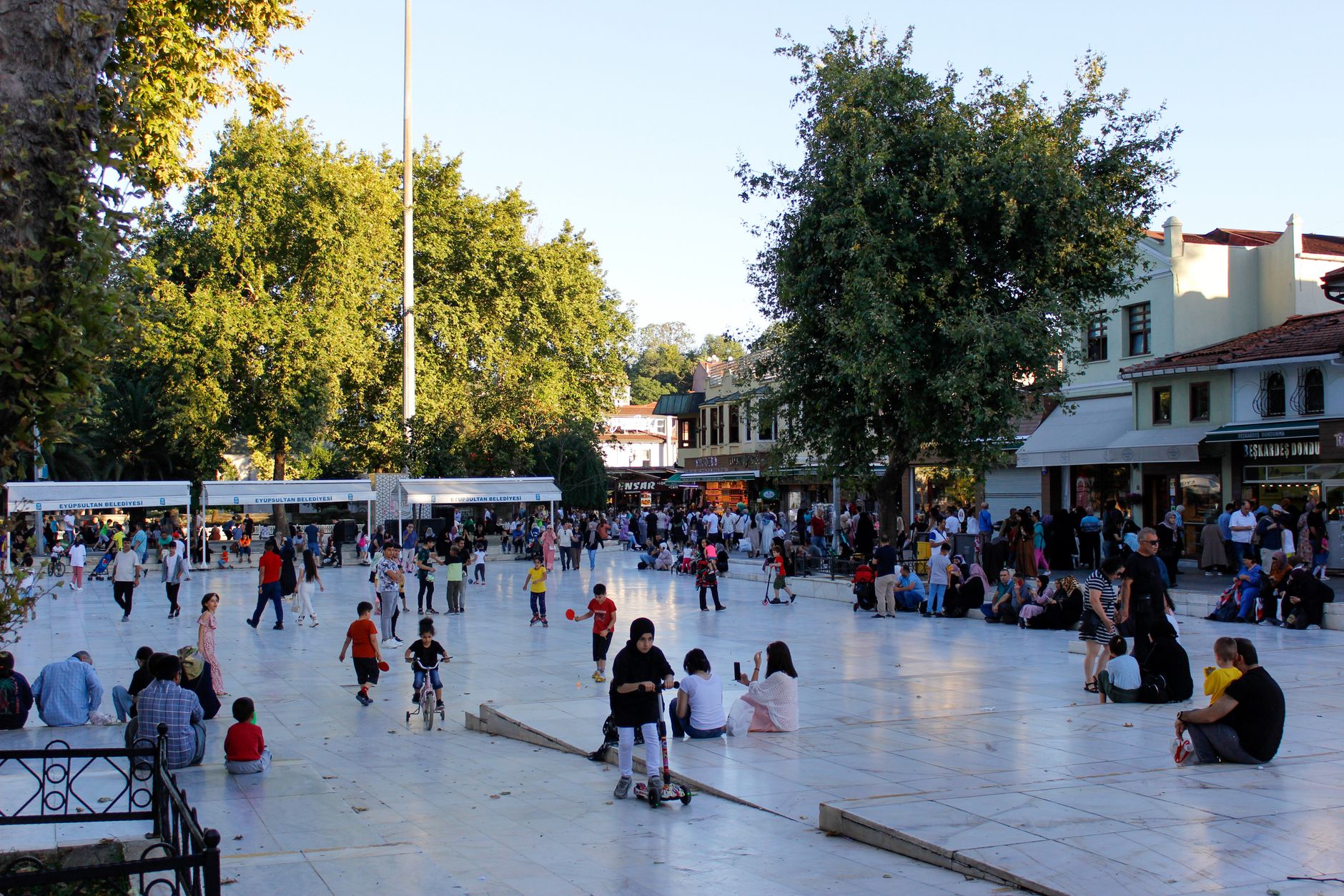 A vibrant city square filled with families, children playing and relaxing under green trees on a sunny day.