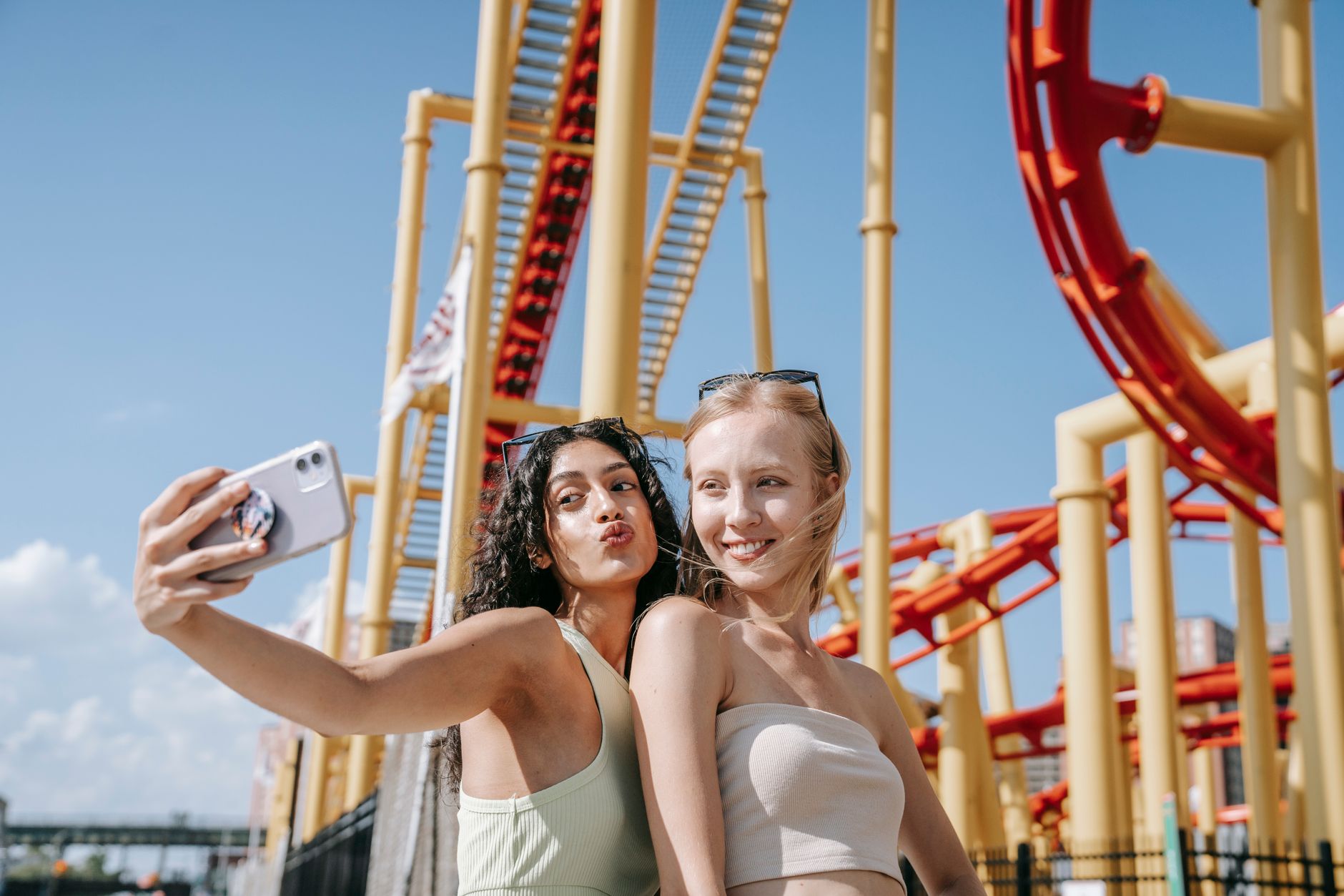 Two young women pose for a selfie in front of a roller coaster on a sunny day.