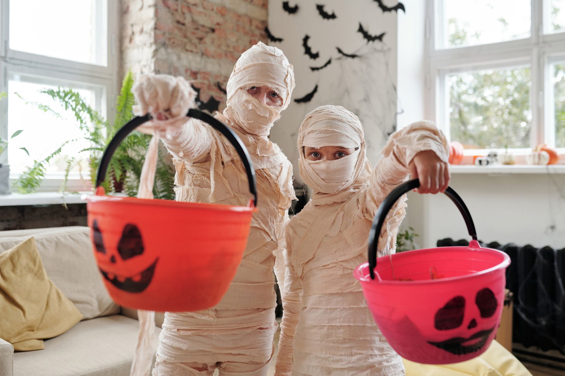 Children dressed as mummies holding Halloween jack-o'-lantern buckets indoors with festive decor.