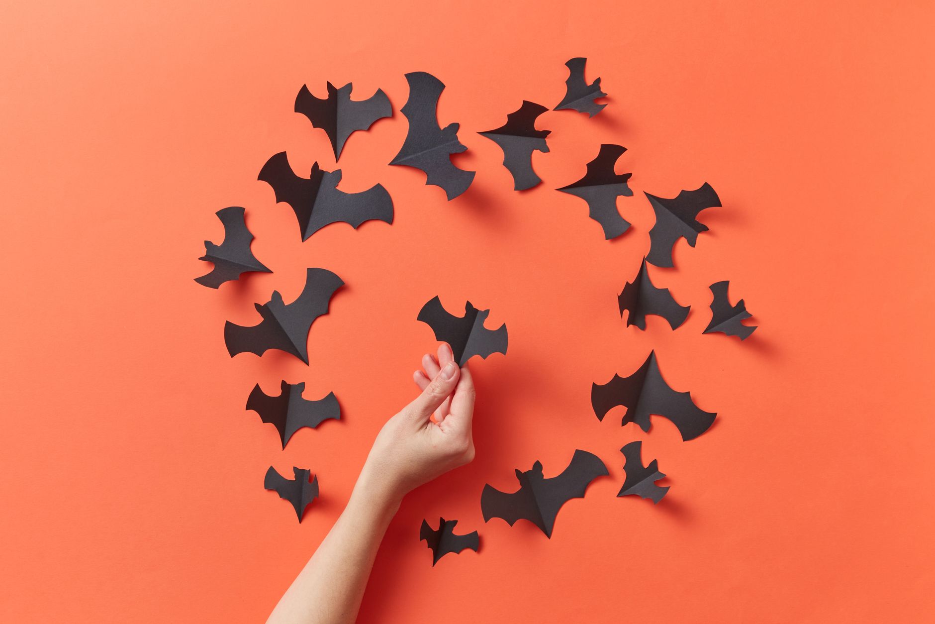A hand arranges paper bats on a vibrant orange background, ideal for Halloween decoration concepts.