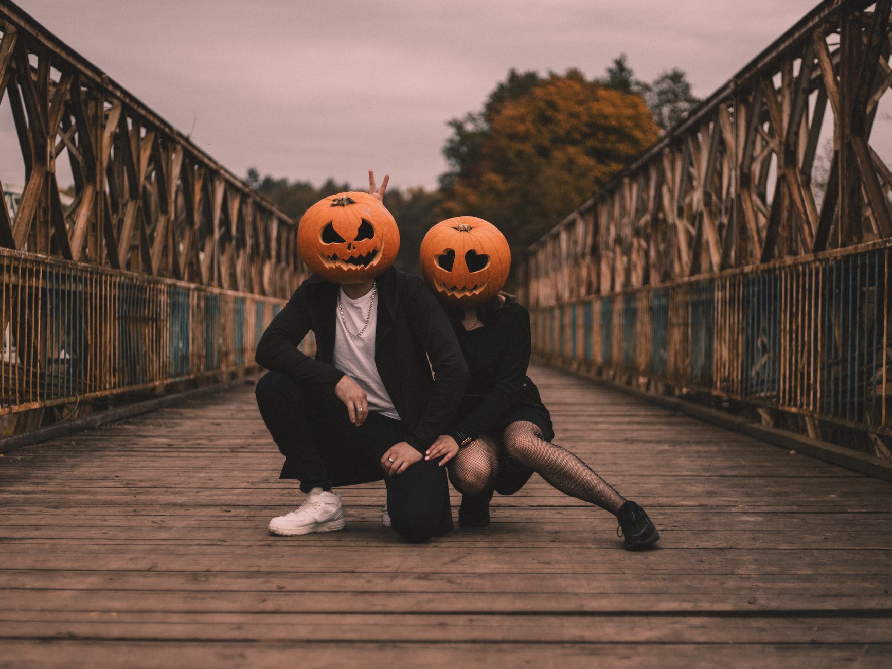 Couple in Halloween costumes with pumpkin heads on an old wooden bridge, adding a spooky charm to the scene.