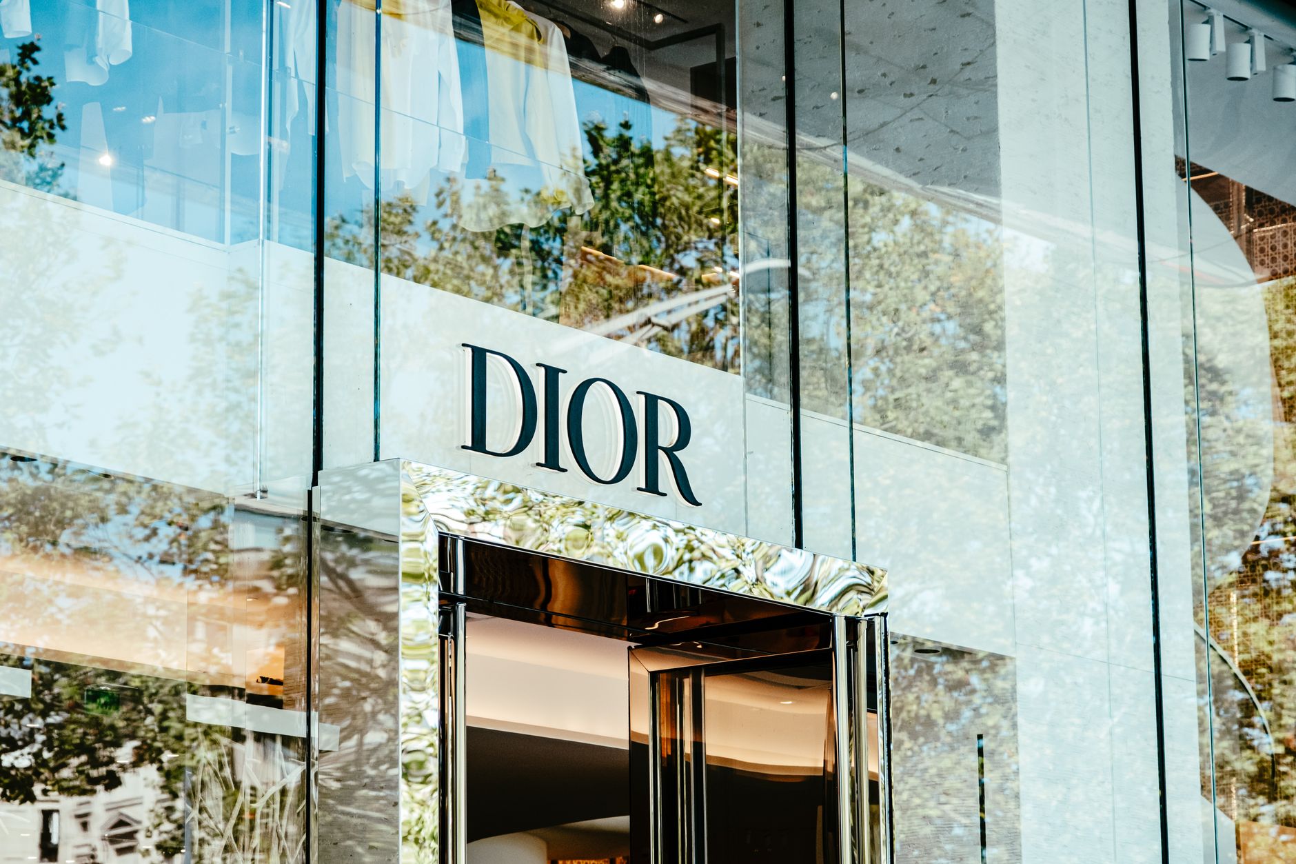 Exterior of a Dior store with glass facade reflecting trees and architecture.