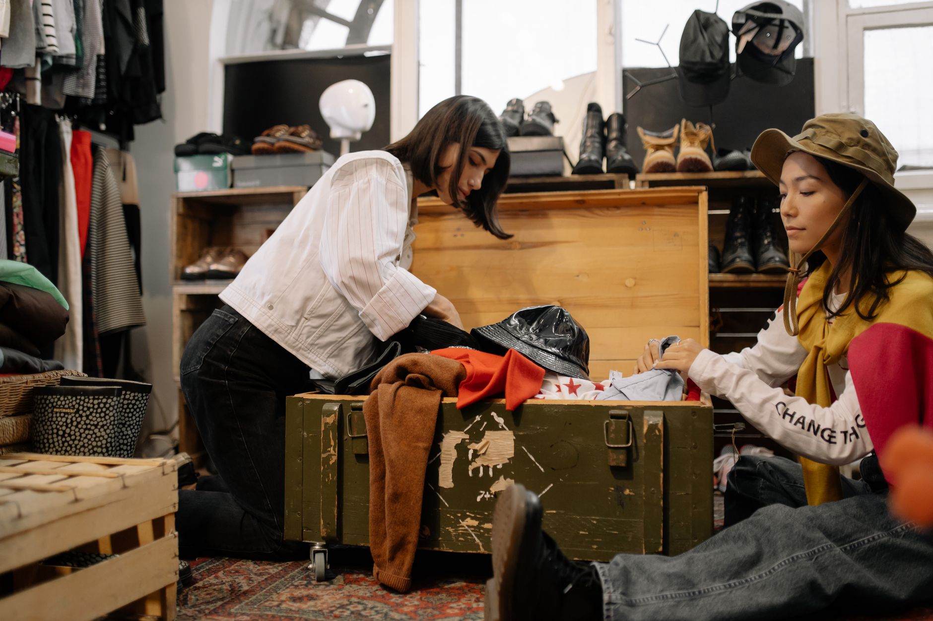 Two women sorting through vintage clothing in a thrift shop, surrounded by shoes and hats.