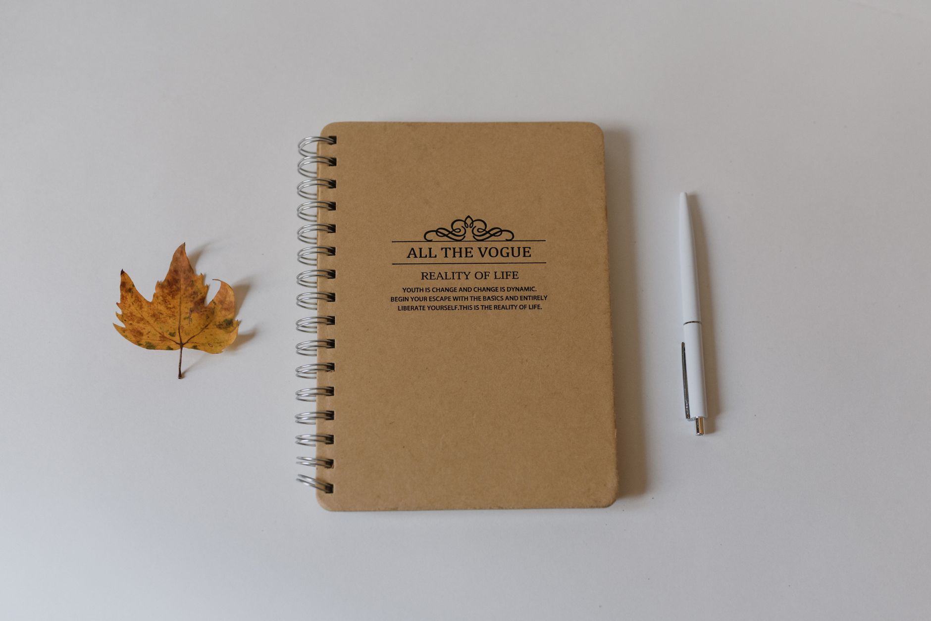 Spiral notebook, dried maple leaf, and a white pen on a clean white background.