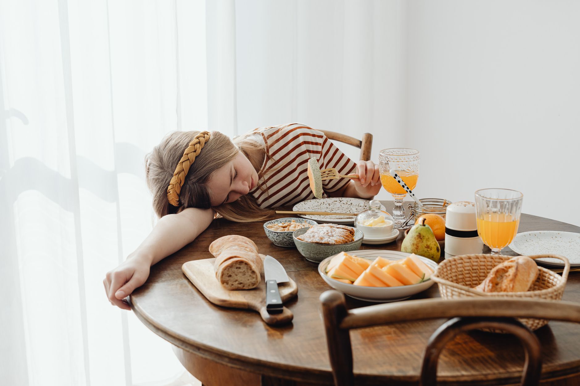 Young girl dozing at breakfast table with diverse food spread, amusing morning scene.
