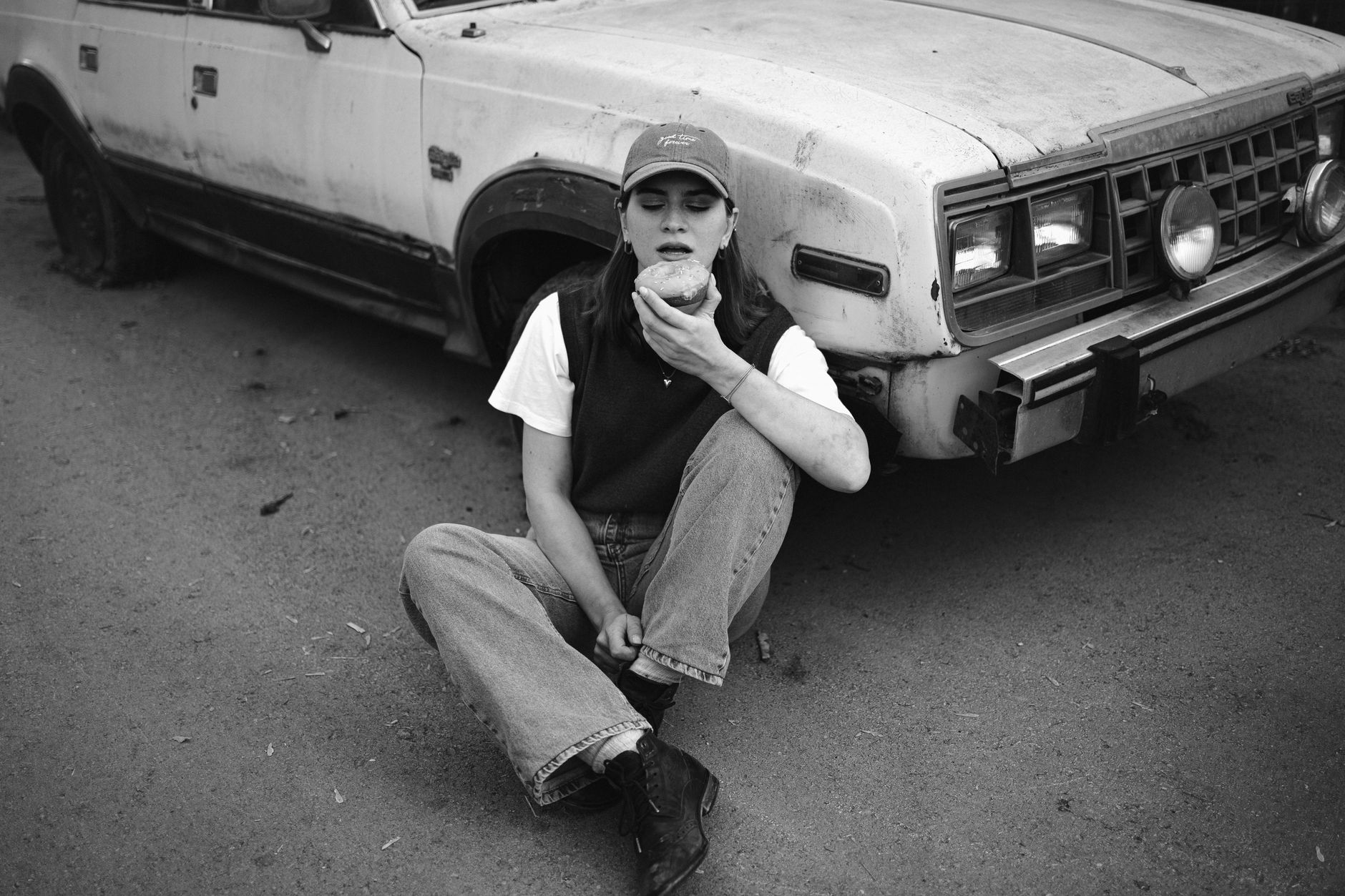 Black and white photo of a woman sitting by a vintage car, savoring a donut.