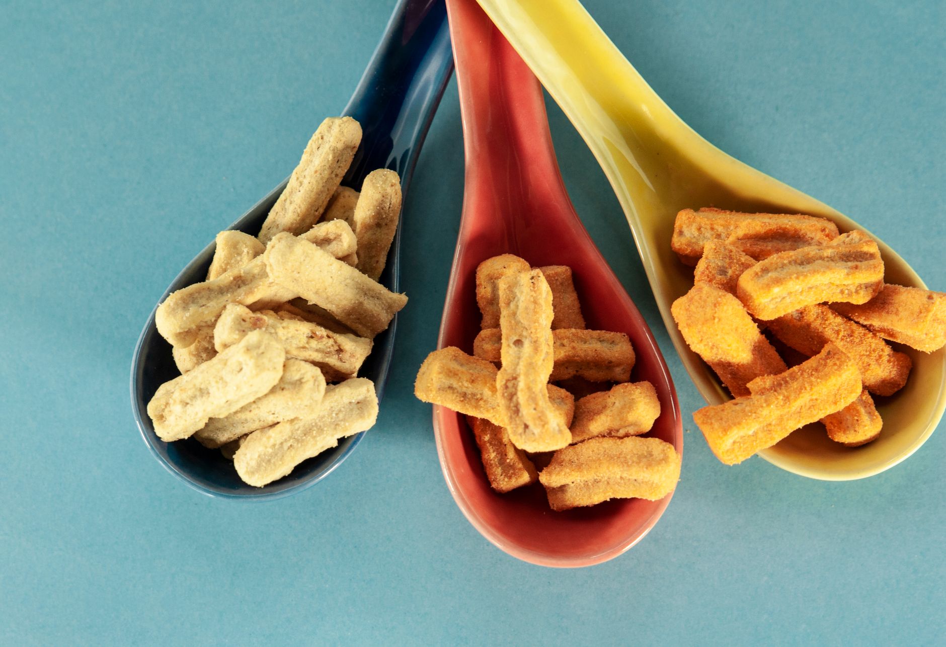 Close-up of three colored spoons holding assorted crunchy snacks on a blue backdrop.