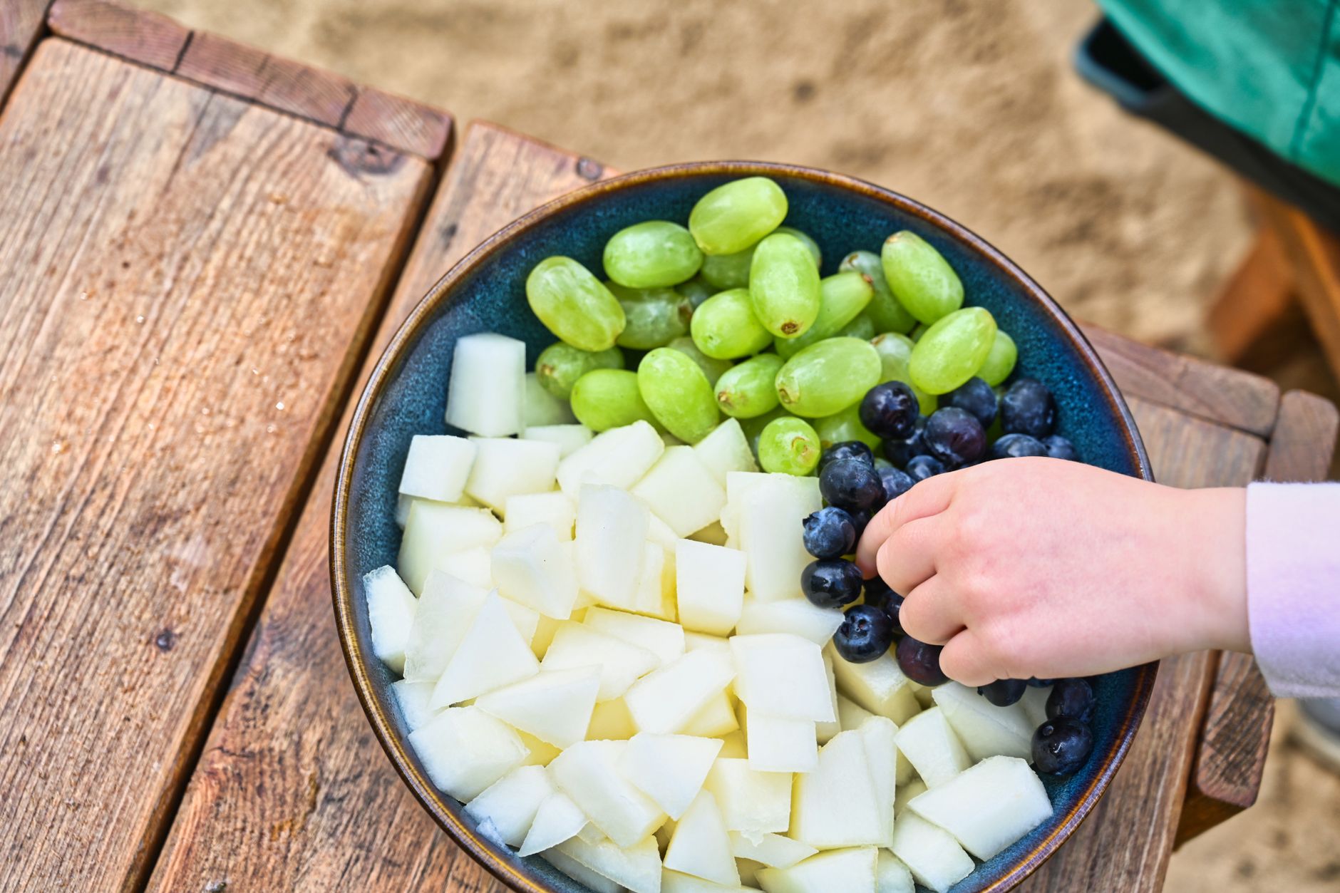 A child's hand reaching for fresh grapes, blueberries, and melon in a bowl on a wooden table.