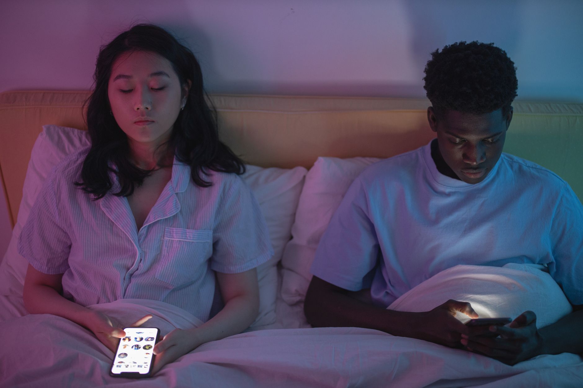 Interracial couple in bed at night using smartphones, illuminated by screen glow, wearing pajamas.