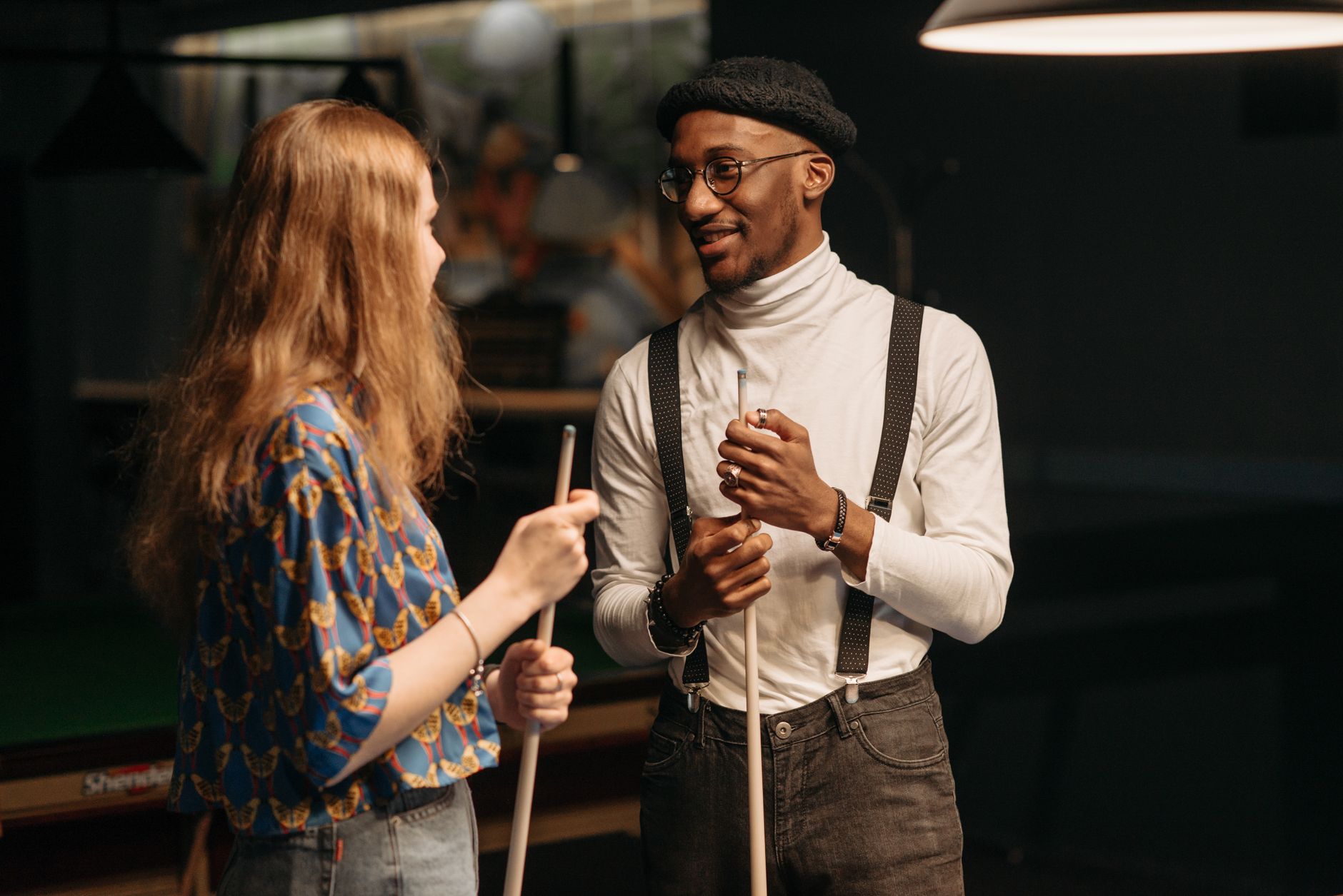 A black man and caucasian woman smiling and conversing while holding cue sticks in a billiards room.
