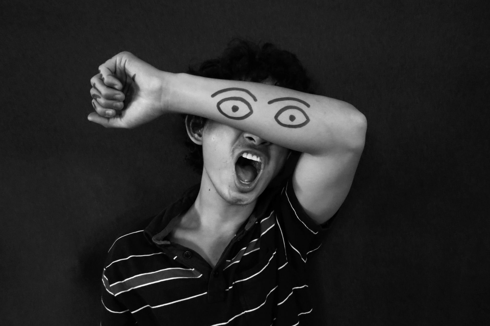 A person covers their eyes with an arm featuring drawn eyes, expressing creativity and emotion.