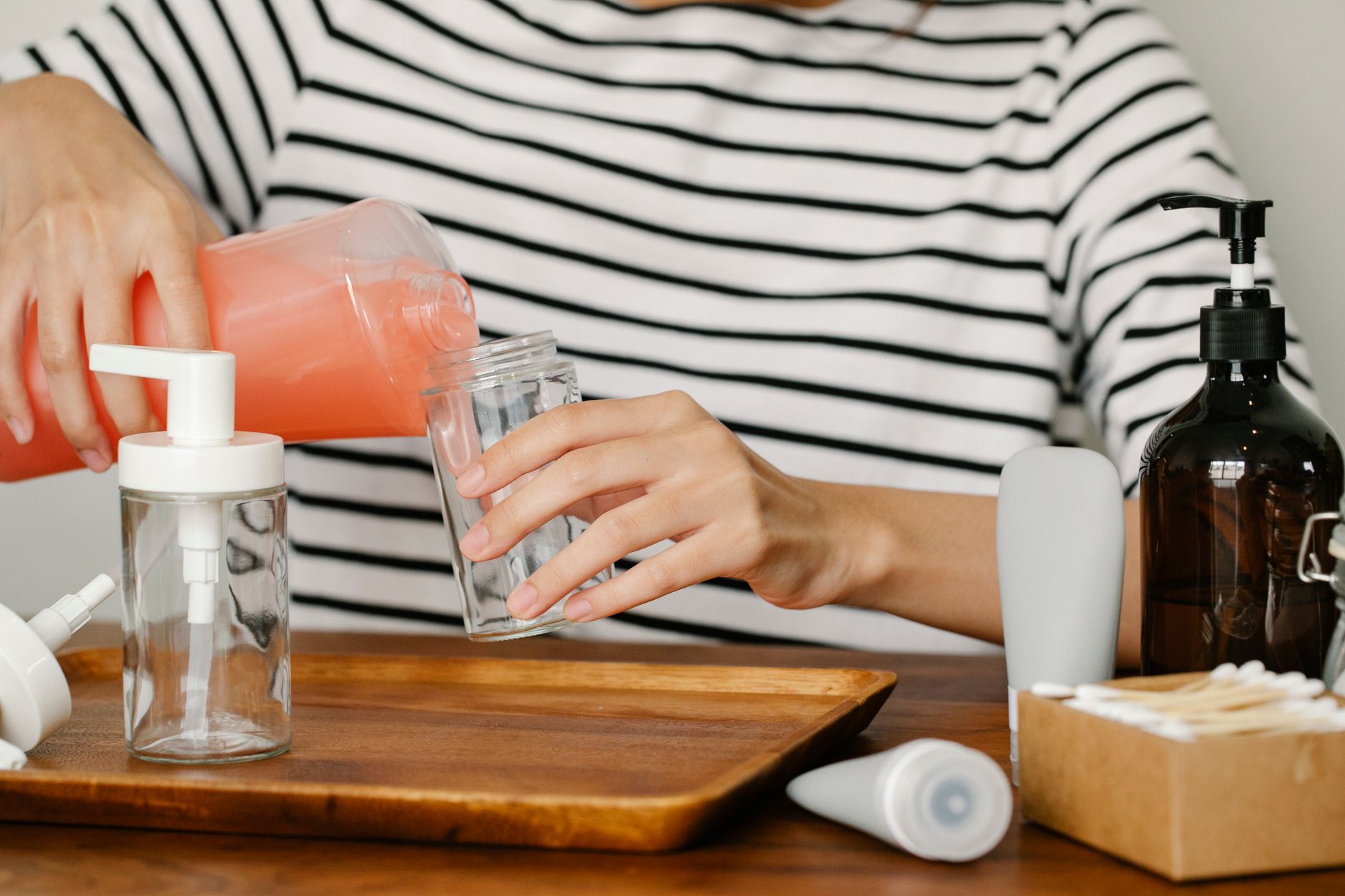 Crop person sitting at table and pouring peach coloured soap from opened soap dispenser into plastic bottle