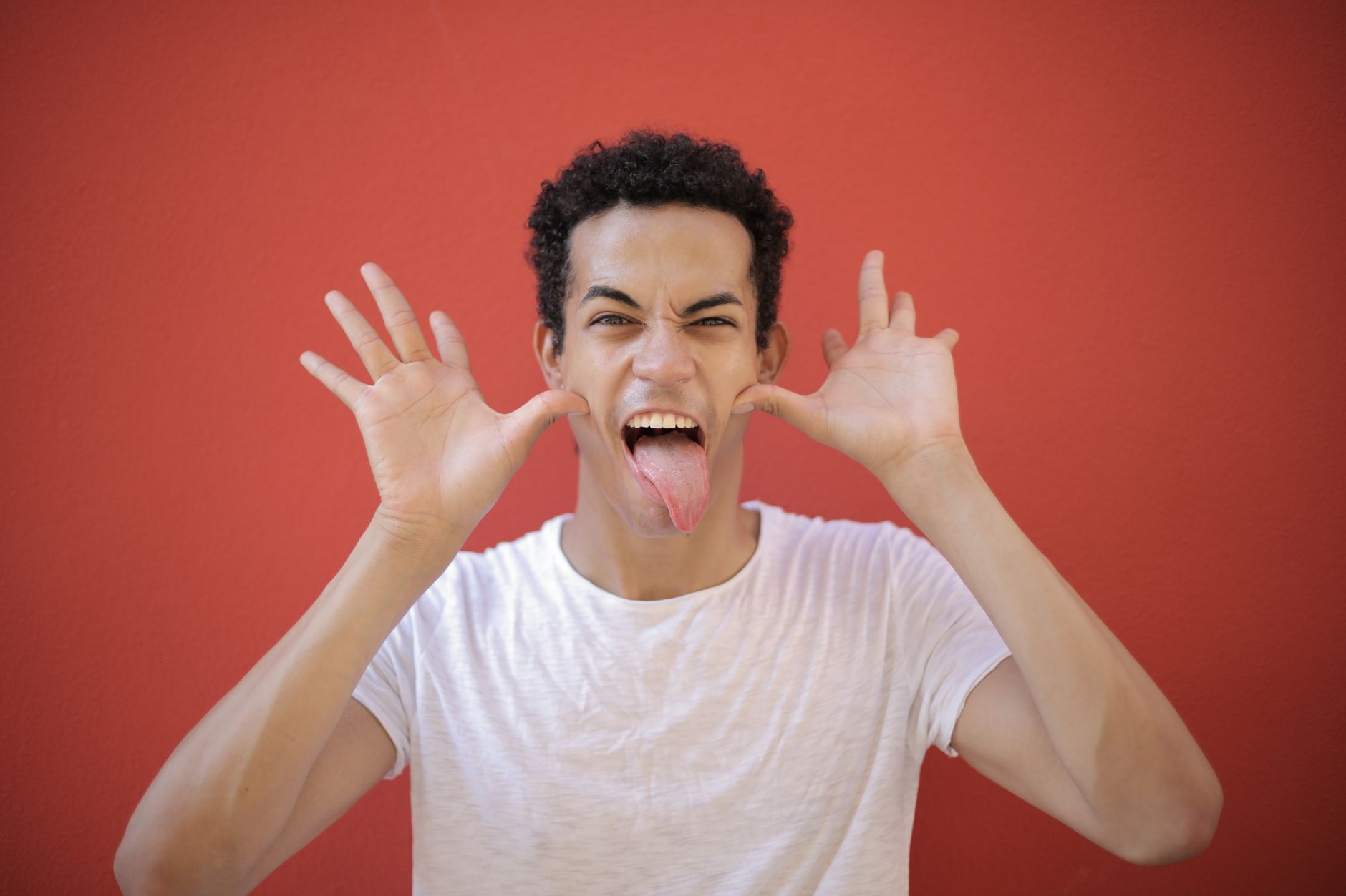 Cheerful young guy with open palms near head sticking out tongue while making grimace against red background