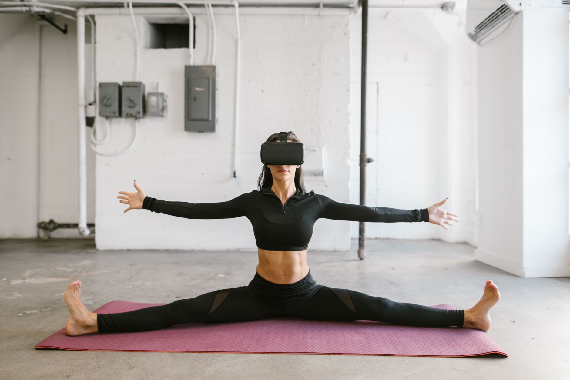 A woman stretches on a yoga mat while wearing a virtual reality headset, indoors.
