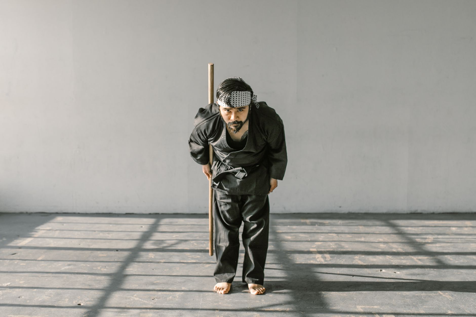 A martial artist in a black gi performs a traditional bow indoors with a wooden staff.