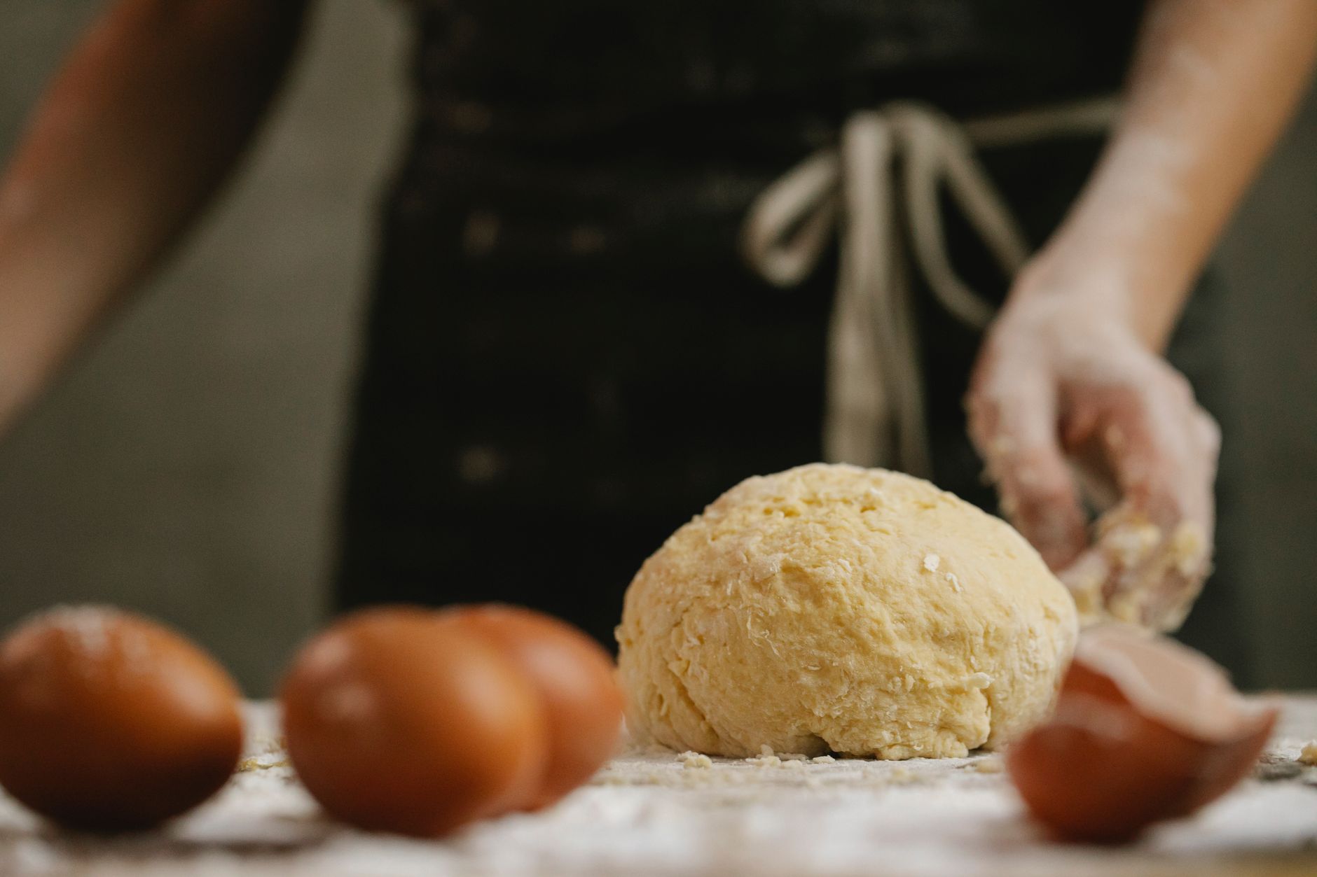 Blurred unrecognizable person in apron standing at table sprinkled with flour and raw dough near scattered eggs in kitchen during cooking process