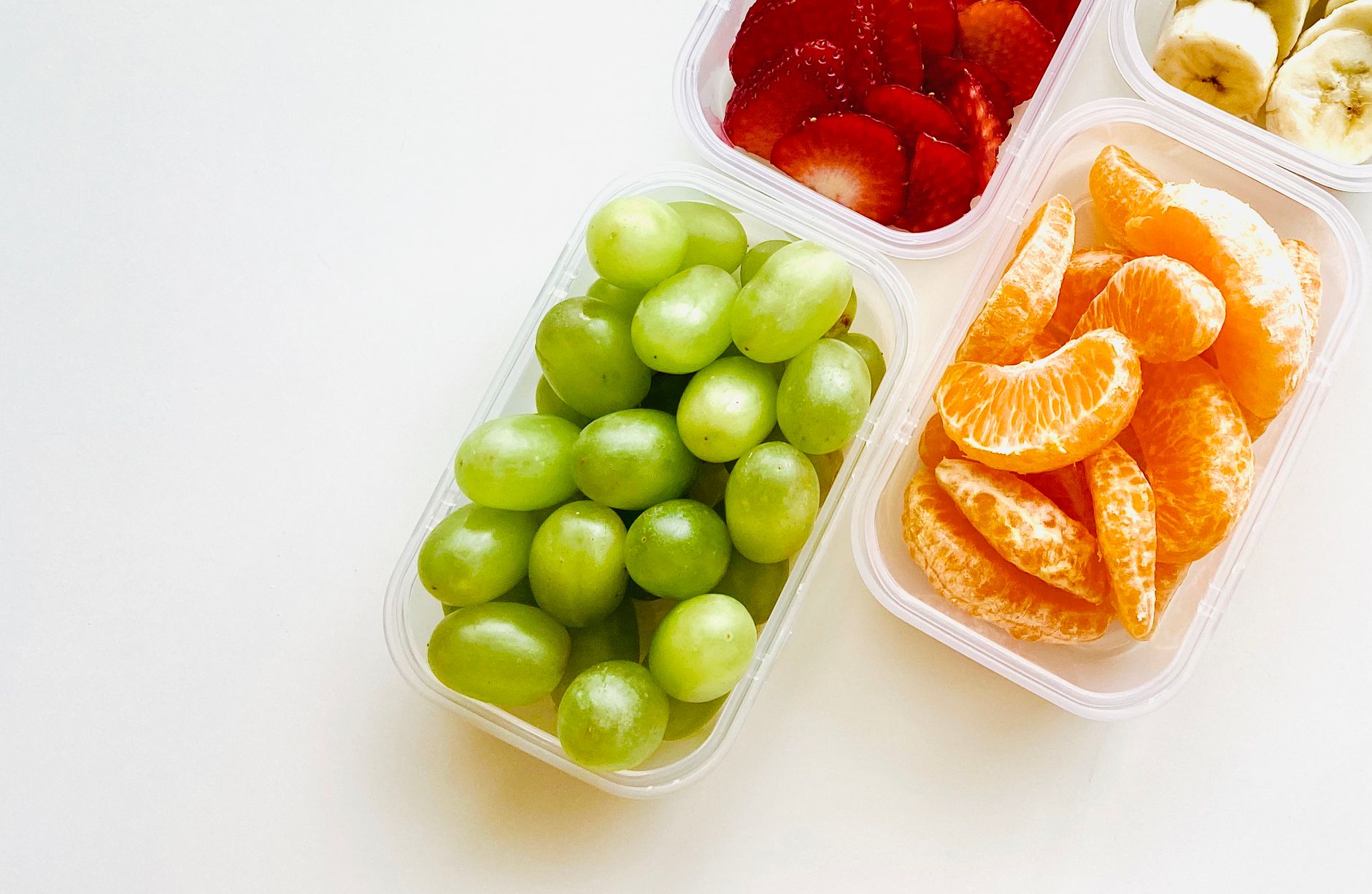 Bright assortment of fresh fruits including grapes, strawberries, orange slices, and bananas in containers.