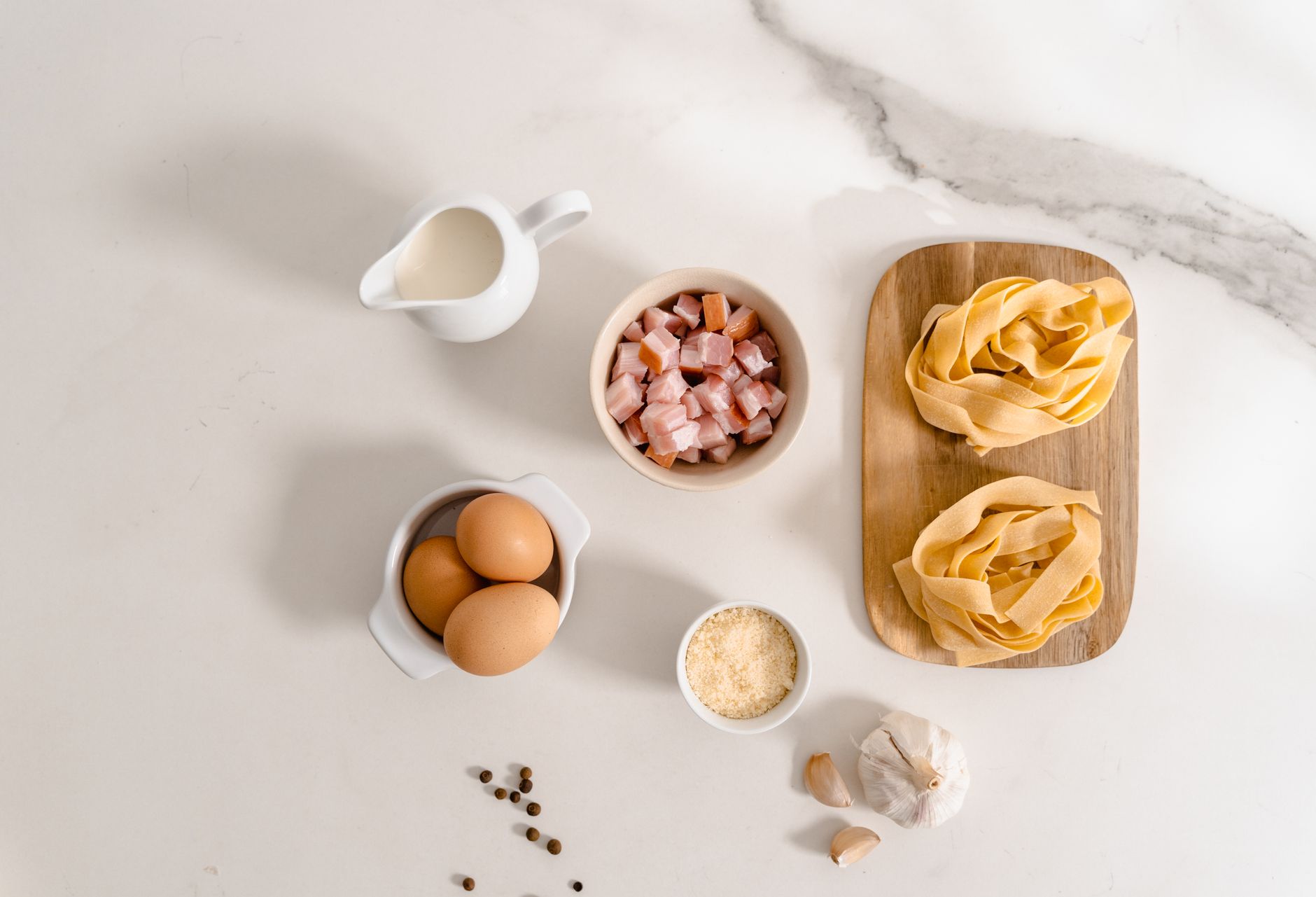 Flat lay of pasta ingredients including eggs, cream, and garlic on marble.
